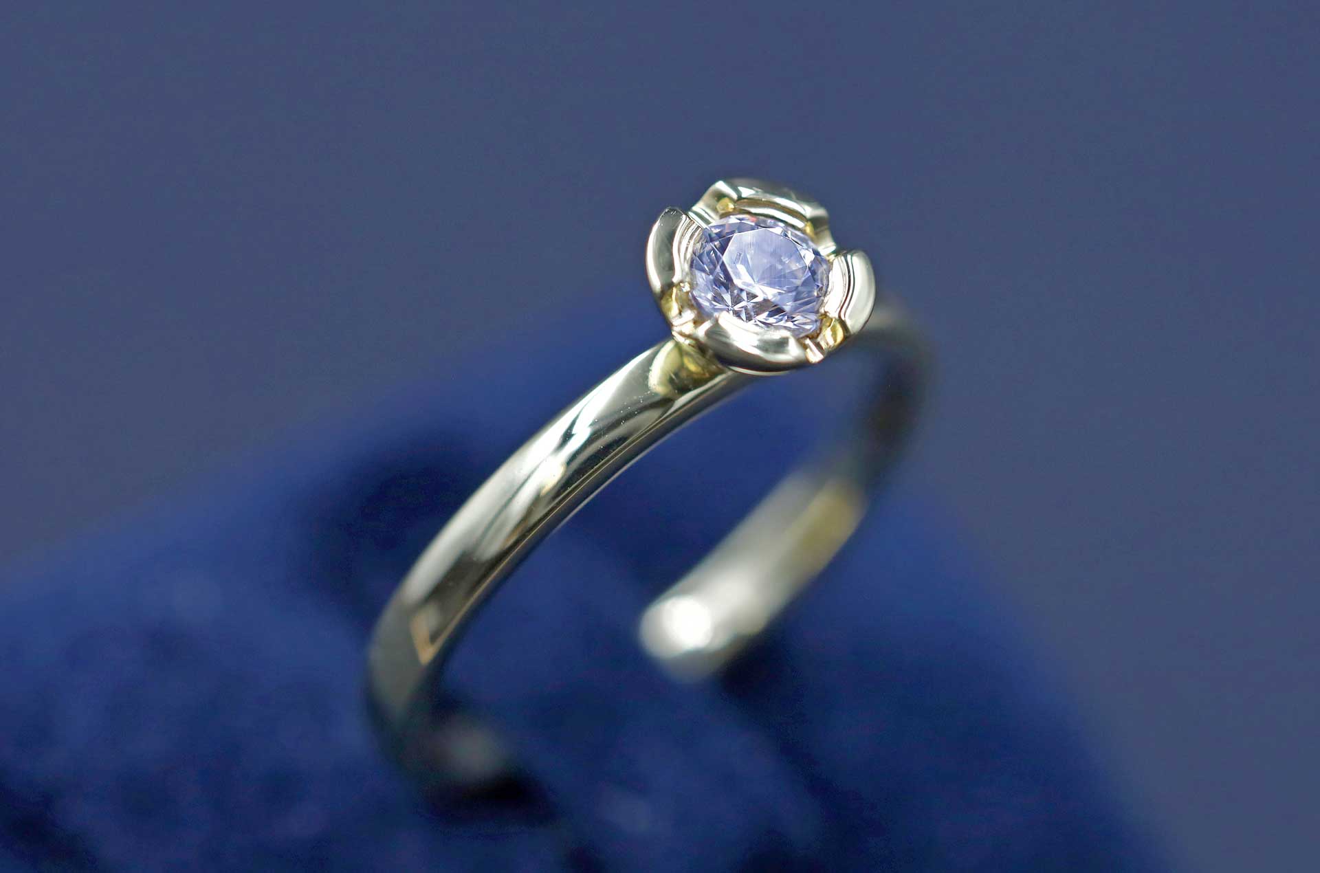 Eleganter Ring aus glänzendem Gelbgold mit einem funkelnd geschliffenen Erinnerungsdiamanten in einer blütenförmigen Fassung auf blauem Samt.