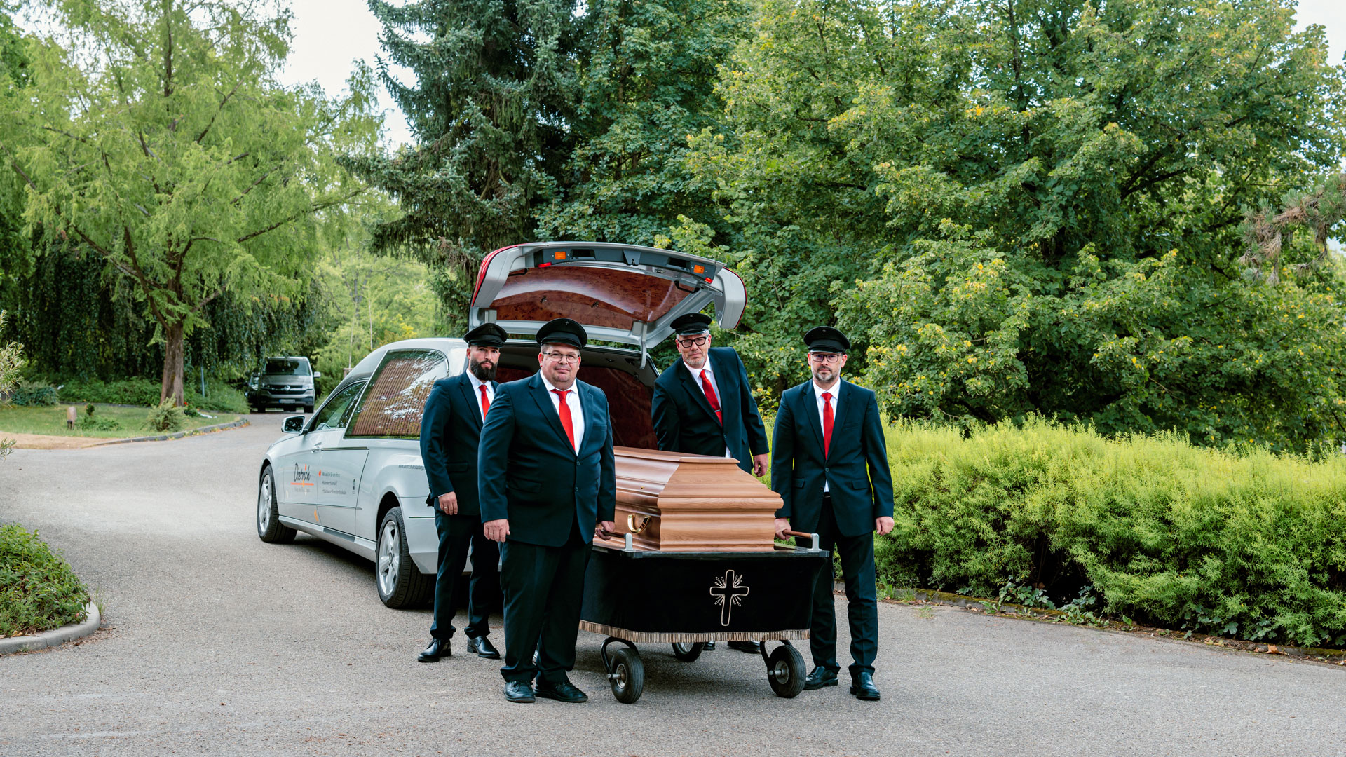 Vier Sargträger des Bestattungshauses Dietrich in dunklen Anzügen und roten Krawatten neben einem Holzsarg und Bestattungswagen auf dem Friedhof Schorndorf.