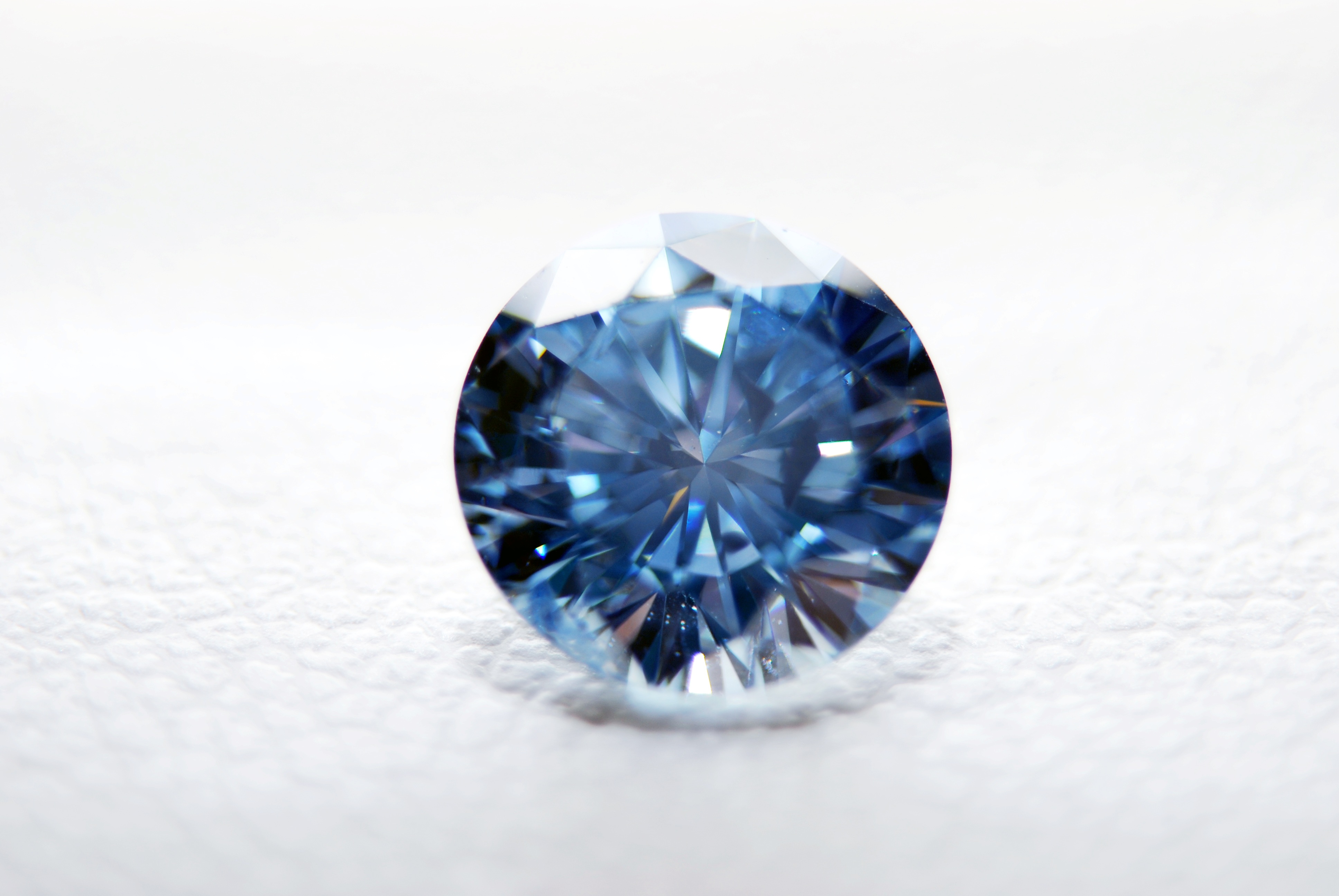 Makroaufnahme eines blau funkelnden Erinnerungsdiamanten im runden Brillantschliff auf einem weißen, leicht strukturierten Hintergrund für eine Diamantbestattung.